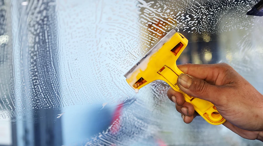 un lavoratore usa un raschietto giallo per rimuovere la colla e lo sporco dal vetro in modo da pulirlo prima di installare una nuova pellicola per vetri